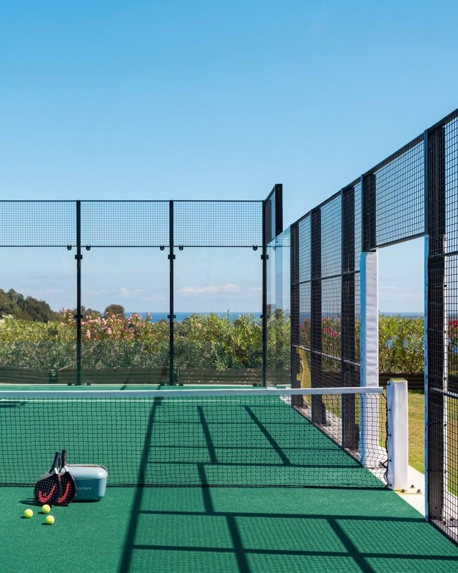 A green padel court beside the net, with two rackets and balls, set in a landscaped garden overlooking the sea